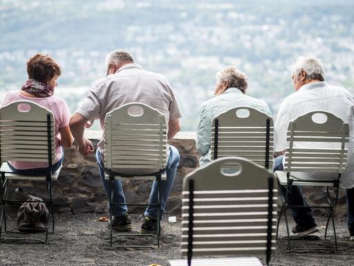 Auf einer Aussichtsplattform stehen vier Stühle auf denen vier ältere Menschen sitzen. Im Hintergrund, in der Unschärfe schaut man auf eine grüne Stadt hinab.