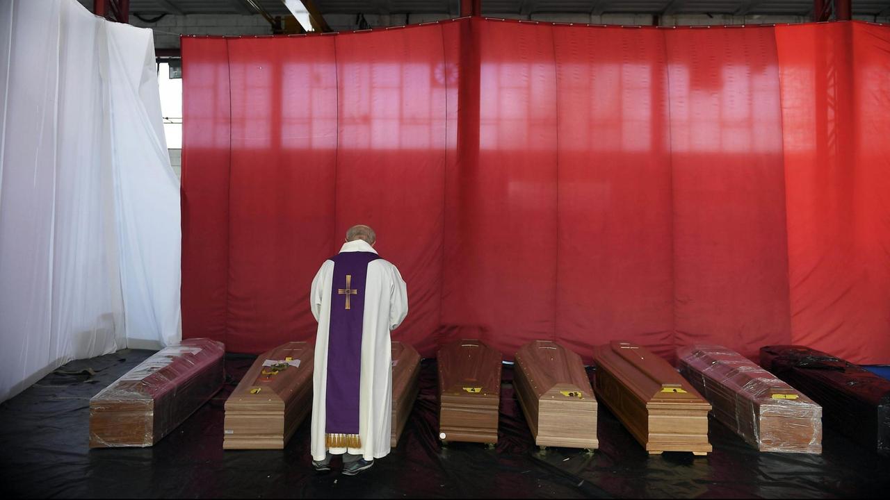 Ein Priester steht vor mehreren aufgereihten Särgen und segnet sie. Ein Priester steht vor mehreren aufgereihten Särgen und segnet sie.