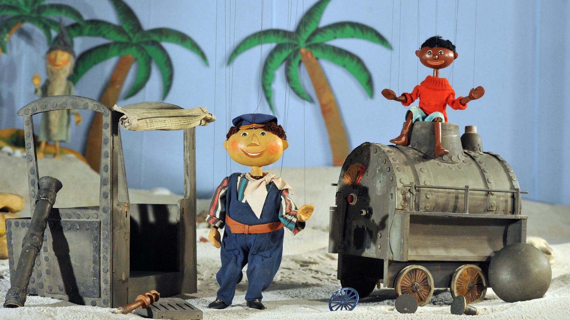 Die Marionetten Scheinriese Tutur, Lukas und Jim Knopf stehen in Augsburg im Puppenkistenmuseum mit der zerstörten Lokomotive Emma in einem Wüstenszenario. Die Marionetten Scheinriese Tutur, Lukas und Jim Knopf stehen in Augsburg im Puppenkistenmuseum mit der zerstörten Lokomotive Emma in einem Wüstenszenario.