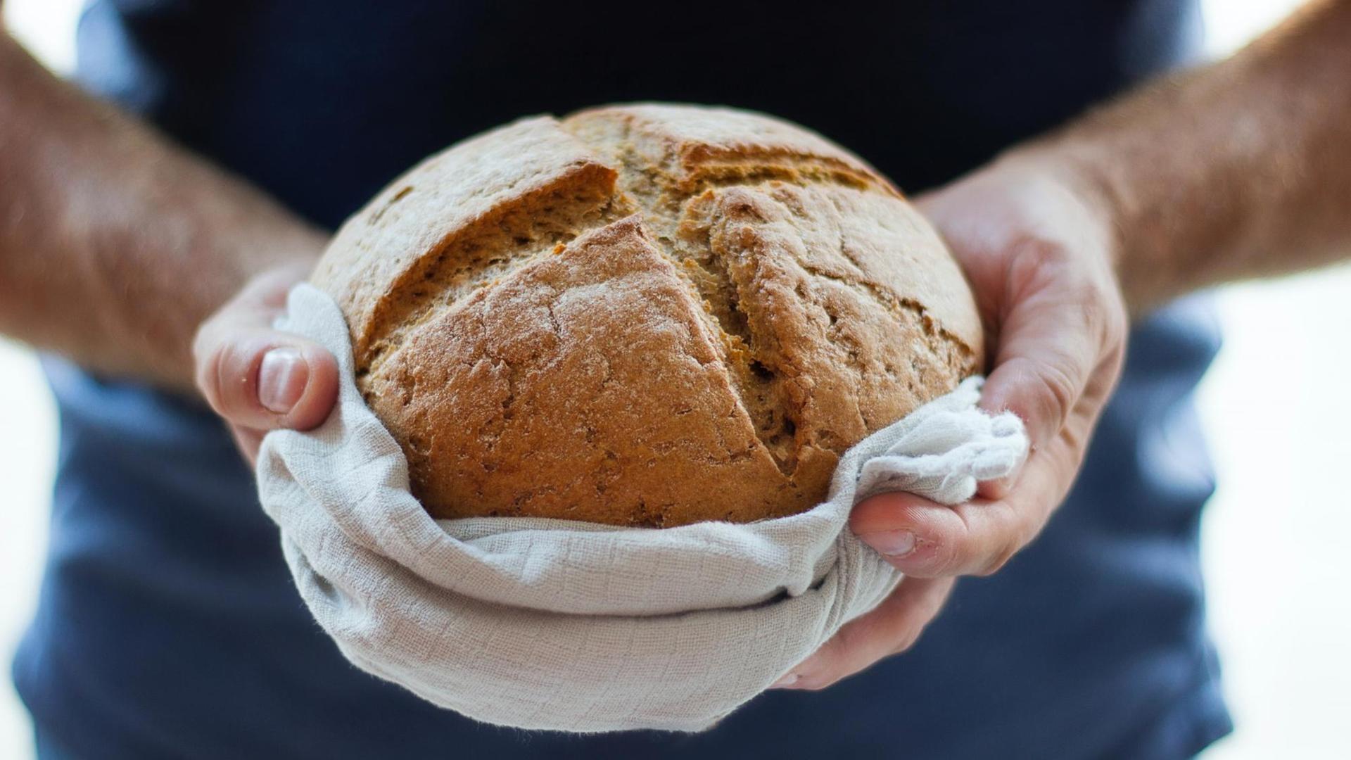 Ein frisch gebackenes Brot in einem Leinentuch von zwei Händen gehalten.