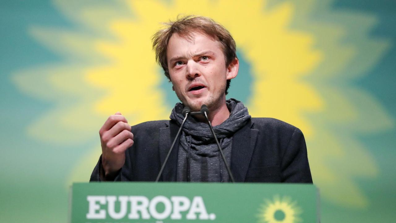 Der junge Grünen-Politiker Erik Marquardt steht an einem Rednerpult und gestikuliert leicht mit der rechten Hand. Im Hintergrund sieht man die Sonnenblume des Grünen-Logos.