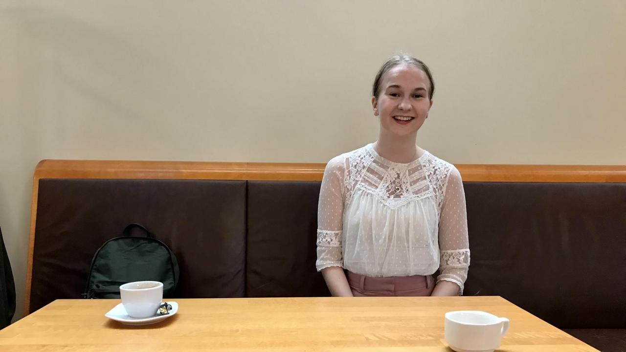 Eine junge Frau mit glatt zurückgebundenem Haar und hochgeschlossener Bluse sitzt vor einem Tisch mit zwei Kaffeetassen. Eine junge Frau mit glatt zurückgebundenem Haar und hochgeschlossener Bluse sitzt vor einem Tisch mit zwei Kaffeetassen.