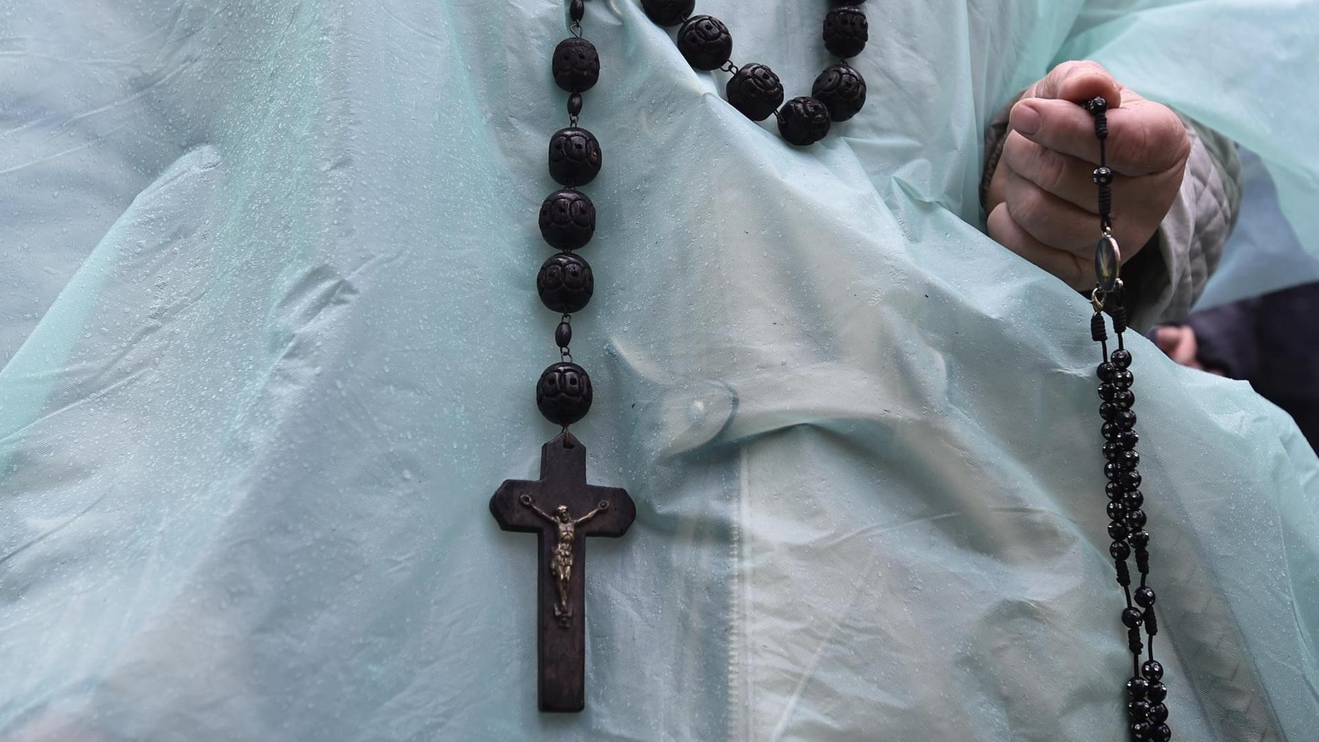 Vor einem weißen Obergewand fällt eine Kette mit einem Kruzifix ab, eine Hand hält einen Rosenkranz. Vor einem weißen Obergewand fällt eine Kette mit einem Kruzifix ab, eine Hand hält einen Rosenkranz.