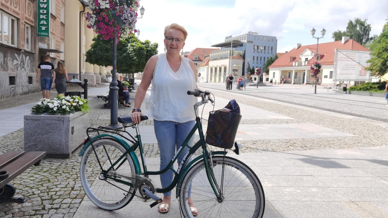 Eine Frau mit kurzen blonden Haaren, Jeans und ärmelloser weißer Bluse schiebt ein Fahrrad.