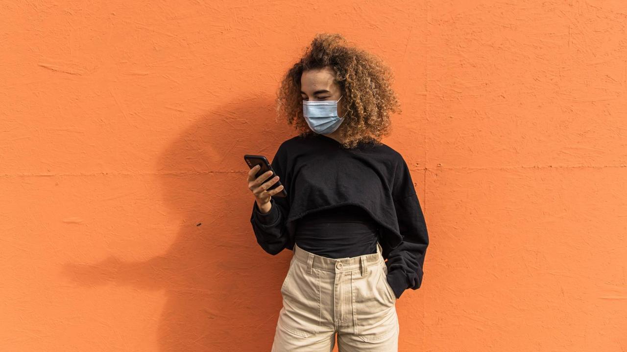 Eine Frau mit Maske steht ihr Smartphone haltend vor einer orange farbenen Wand