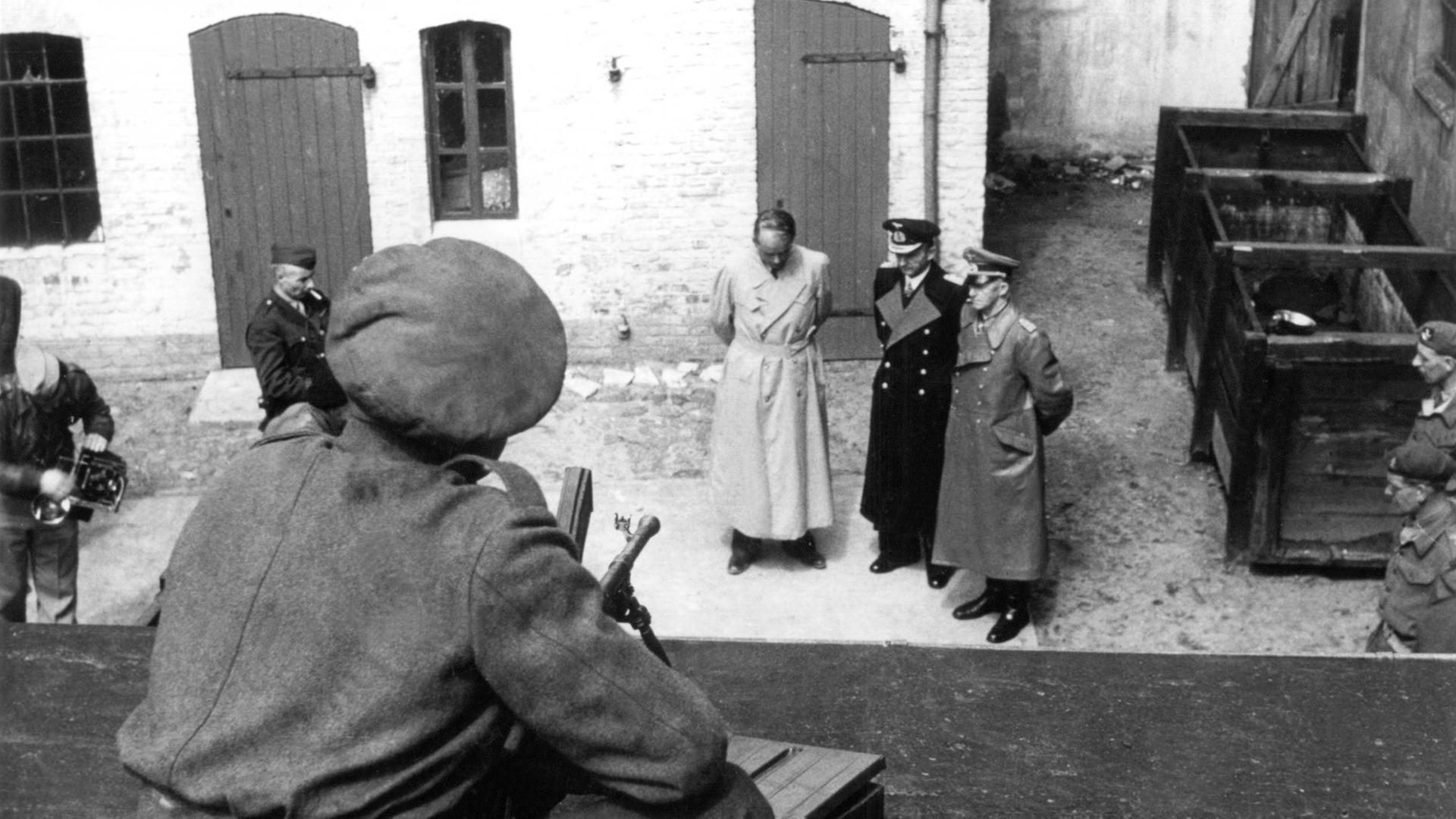 Albert Speer, Karl Dönitz und Alfred Jodl stehen in Trenchoats und Uniformen in einem Hof, umringt von britischen Soldaten, von denen zwei eine Kamera aufstellen. Aufgenommen ist das Bild von einem Dach aus, auf dem ein Soldat mit einem Gewehr im Anschlag kauert.