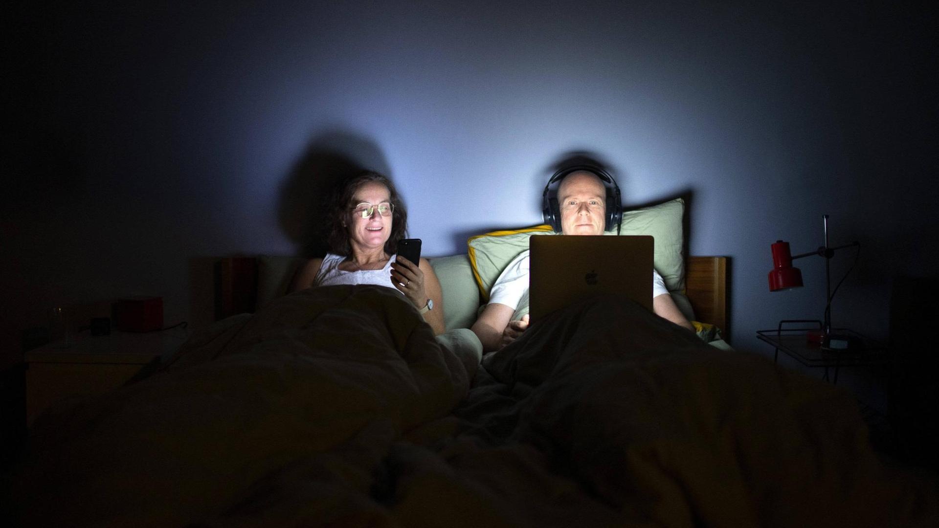 Paar liegt im Bett, die Frau schaut auf ihr Smartphone, während der Mann Kopfhörer aufhat und auf sein Laptop starrt.