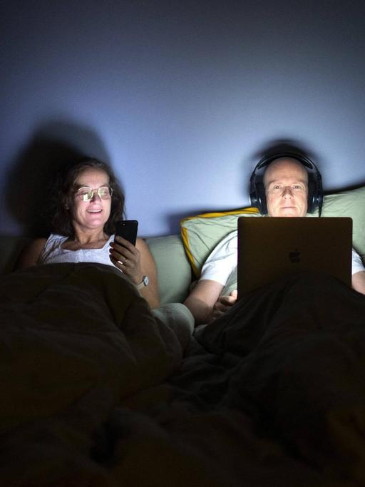 Paar liegt im Bett, die Frau schaut auf ihr Smartphone, während der Mann Kopfhörer aufhat und auf sein Laptop starrt.