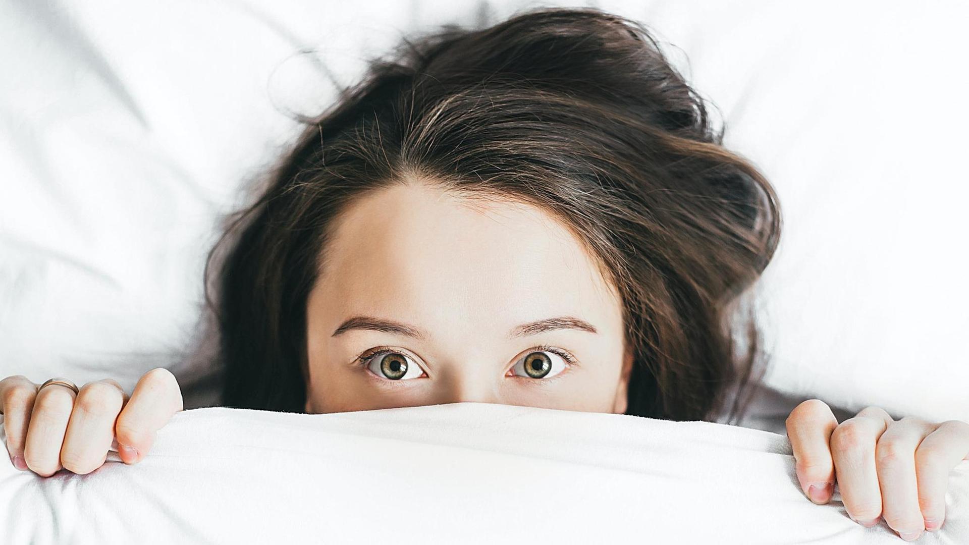Eine Person zieht die weiße Bettdecke soweit über das Gesicht, dass nur noch die Augen und die Stirn zu erkennen sind. Eine Person zieht die weiße Bettdecke soweit über das Gesicht, dass nur noch die Augen und die Stirn zu erkennen sind.