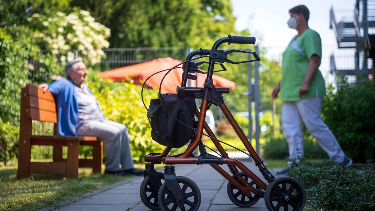 Ein Rollator steht nahe einer Pflegefachkraft und einer Bewohnerin des Seniorenheims "Mein Zuhause Nienburg" im Freien. Die Seniorin sitzt auf einer Parkbank, die Pflegerin steht vor ihr und trägt einen Mund-Nase-Schutz.