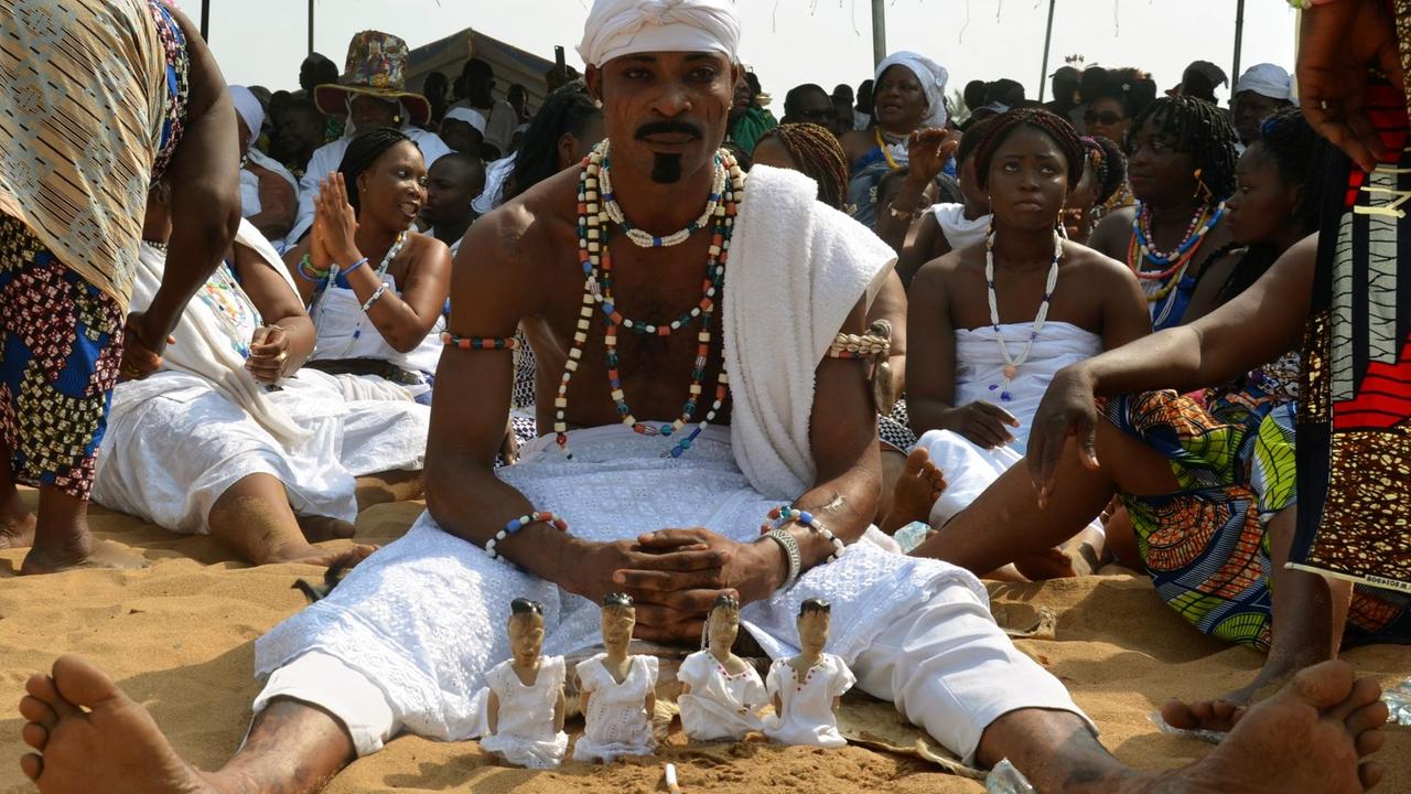 Ein Mann sitzt am Strand von Benin mit Holzpüppchen. Diese symbolisieren, dass ein Zwilling verloren wurde und erinnern daran.