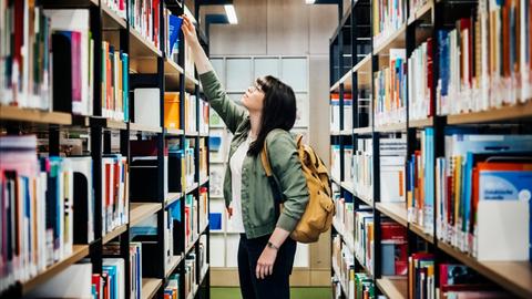 Eine junge Studentin nimmt ein Buch aus dem Regal einer Uni Bibliothek.