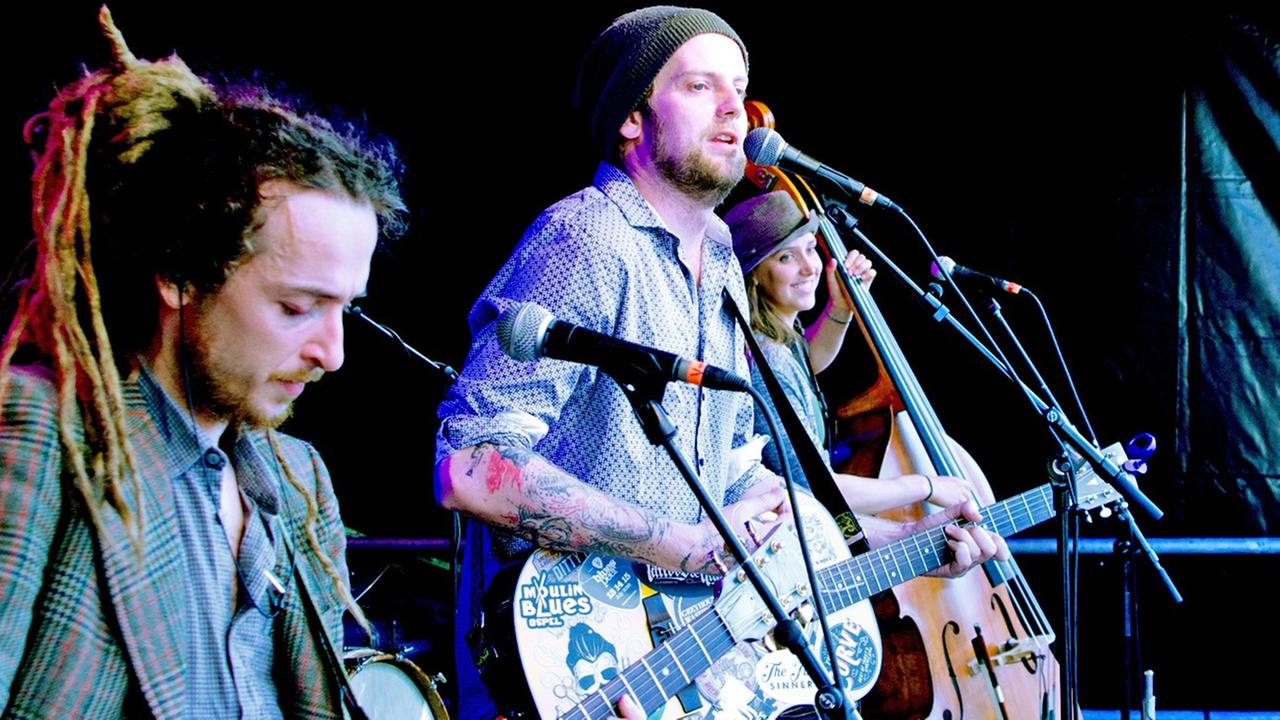 Im Vordergrund stehen zwei Männer an Gitarren, dahinter spielt eine Frau Kontrabaß. Im Vordergrund stehen zwei Männer an Gitarren, dahinter spielt eine Frau Kontrabaß.
