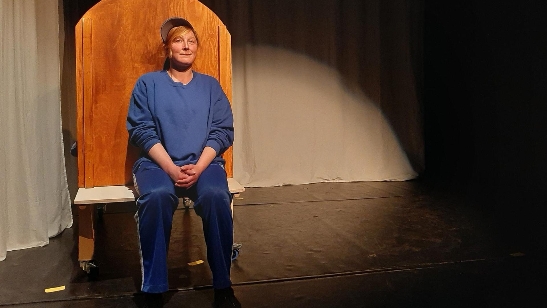 Die ehemals abhänge Schauspielerin Kathi sitzt auf einer hölzernen Bank auf einer Bühne und lächelt verschmitzt in die Kamera. Sie ist komplett in blau gekleidet und trägt eine Cap unter der ihr blonden Haare herausschauen. Die ehemals abhänge Schauspielerin Kathi sitzt auf einer hölzernen Bank auf einer Bühne und lächelt verschmitzt in die Kamera. Sie ist komplett in blau gekleidet und trägt eine Cap unter der ihr blonden Haare herausschauen.