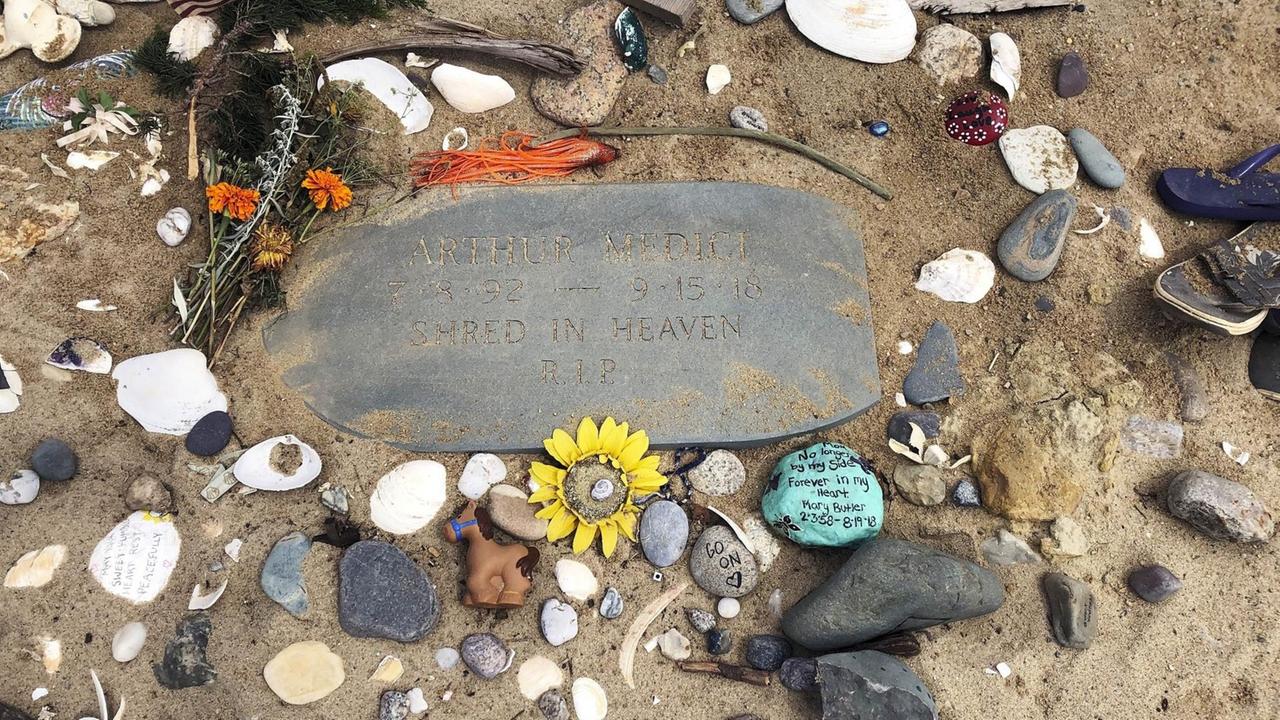 Um eine Gedenkplatte aus Stein im Sand liegen viele weitere kleine Steine, Muscheln und Blumen. 