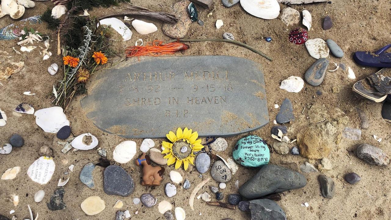 Um eine Gedenkplatte aus Stein im Sand liegen viele weitere kleine Steine, Muscheln und Blumen. Um eine Gedenkplatte aus Stein im Sand liegen viele weitere kleine Steine, Muscheln und Blumen.