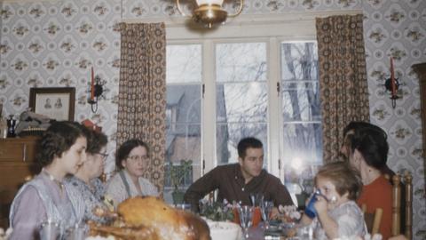 Farb-Foto einer Familie. Zu sehen sind sieben Familienmitglieder verschiedenen Alterns, die an einem gedeckten Tisch sitzen, auf dem ein knuspriger Braten steht. Das Foto ist alt und leicht unscharf.