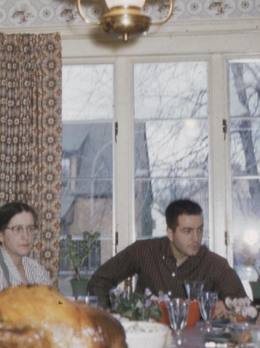 Farb-Foto einer Familie. Zu sehen sind sieben Familienmitglieder verschiedenen Alterns, die an einem gedeckten Tisch sitzen, auf dem ein knuspriger Braten steht. Das Foto ist alt und leicht unscharf.