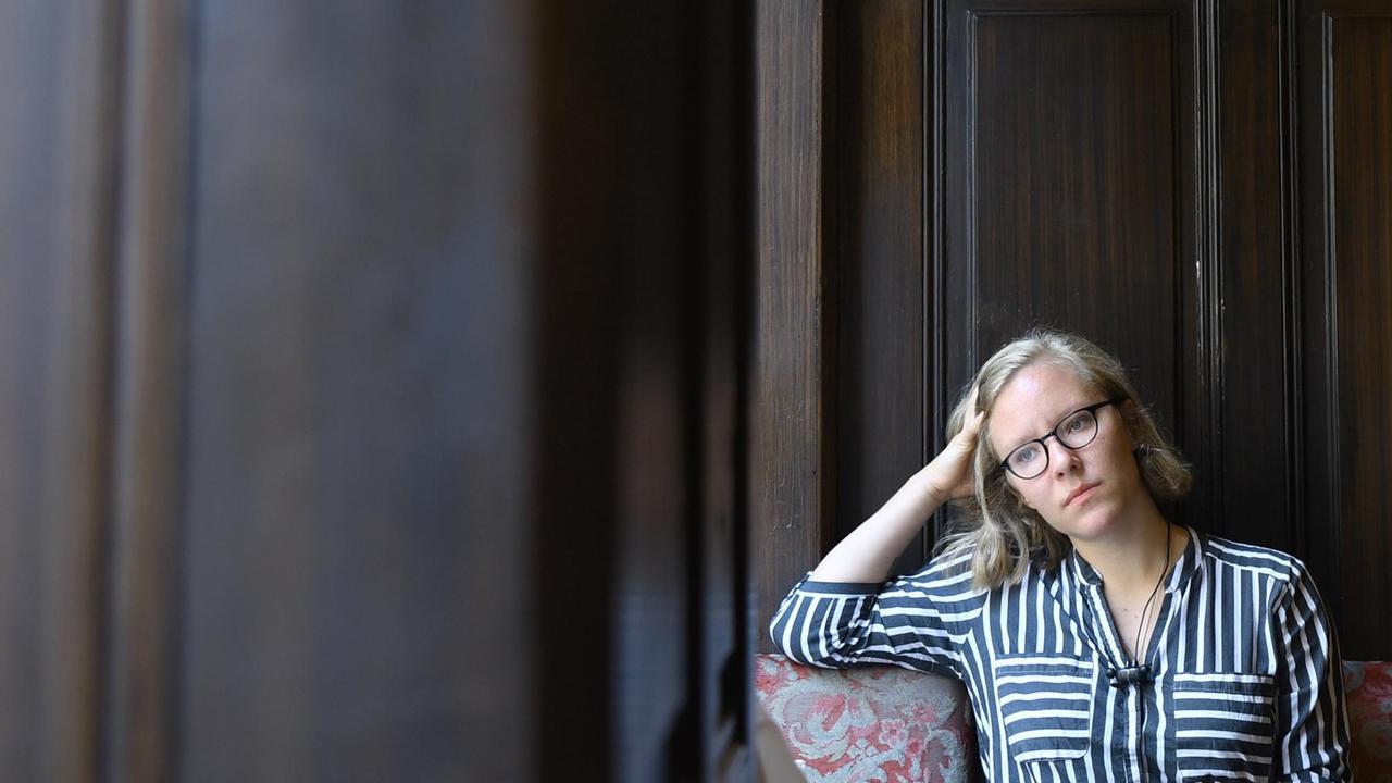 Das Bild zeigt die die österreichische Schriftstellerin Raphaela Edelbauer, die vor einer dunklen Holzwand sitzt. Das Bild zeigt die die österreichische Schriftstellerin Raphaela Edelbauer, die vor einer dunklen Holzwand sitzt.