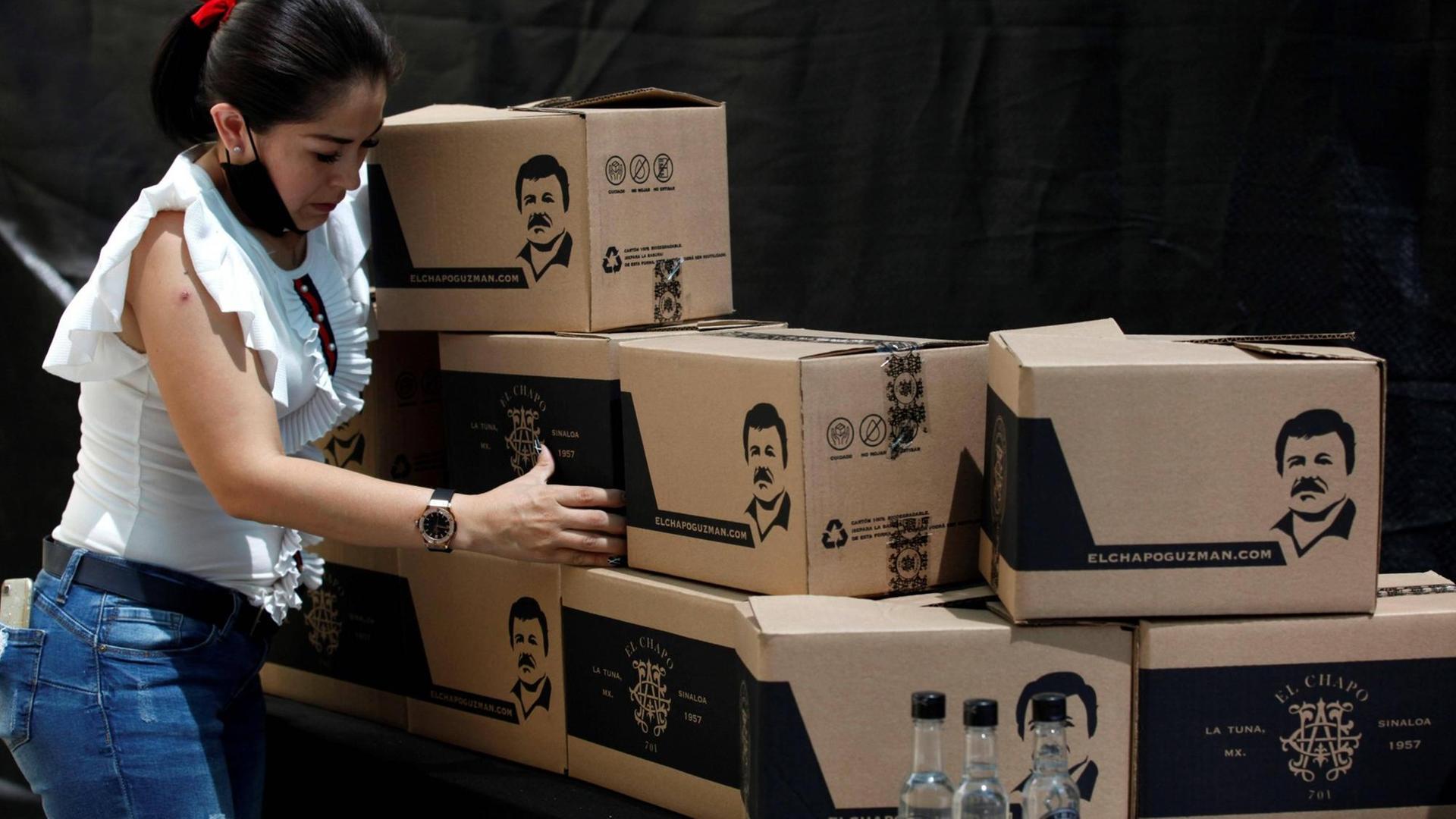 Eine Frau mit Atemschutzmaske um den Hals stapelt Kisten, die mit einer Grafik von Joaquin El Chapo Guzman versehen sind. Eine Frau mit Atemschutzmaske um den Hals stapelt Kisten, die mit einer Grafik von Joaquin El Chapo Guzman versehen sind.