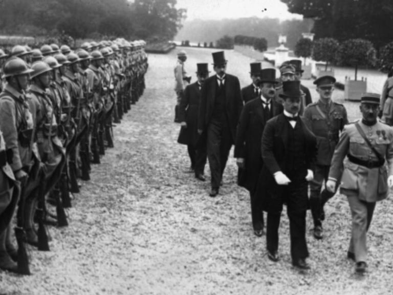 Ankunft der ungarischen Delegation, unter Führung von Dr. Benard, begleitet von Colonel Henry. Männer in Anzügen laufen an Soldatenreihen vorbei.