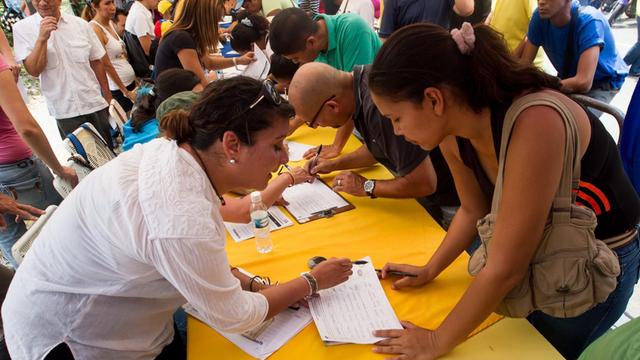Menschen beugen sich über einen gelben Tisch, um sich in Unterschriftenlisten einzutragen.
