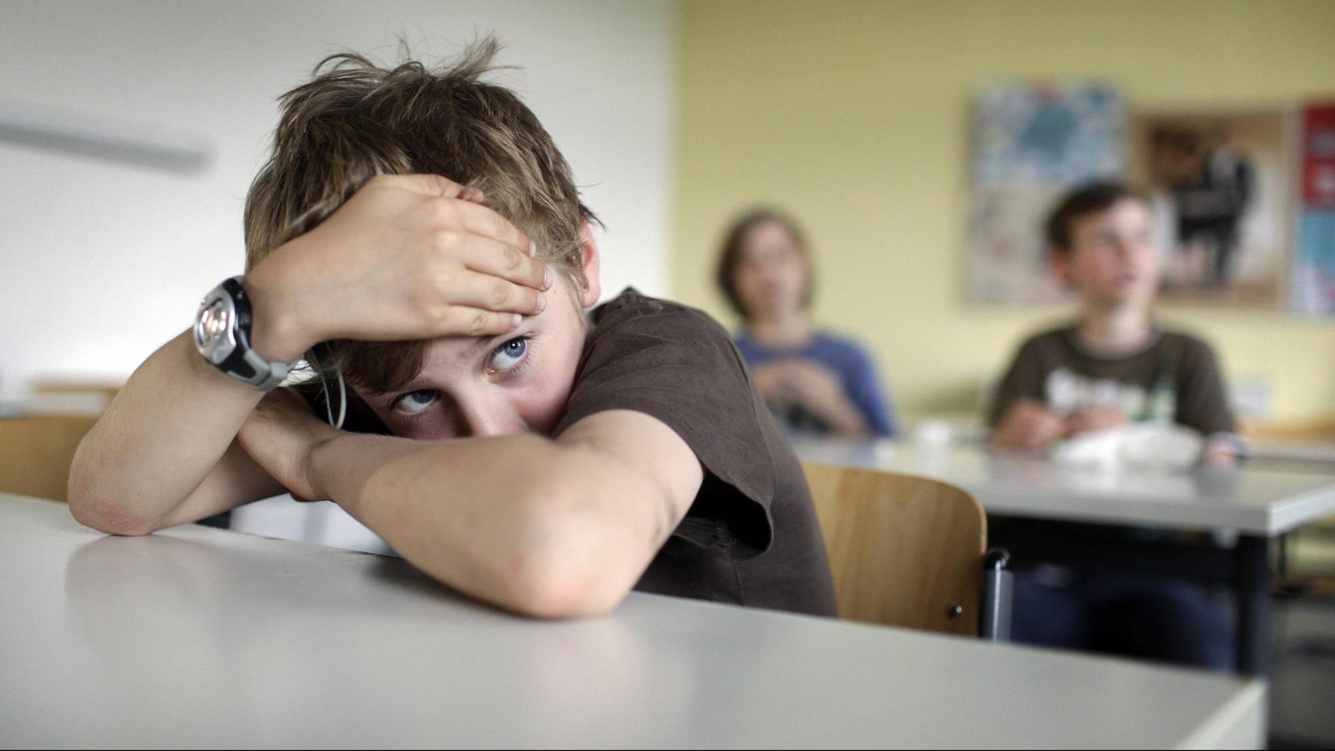 Ein Junge mit dunklem T-Shirt und zerzausten Haaren sitzt im Klassenzimmer und hat den Kopf auf die verschränkten Arme gestützt. Ein Junge mit dunklem T-Shirt und zerzausten Haaren sitzt im Klassenzimmer und hat den Kopf auf die verschränkten Arme gestützt.