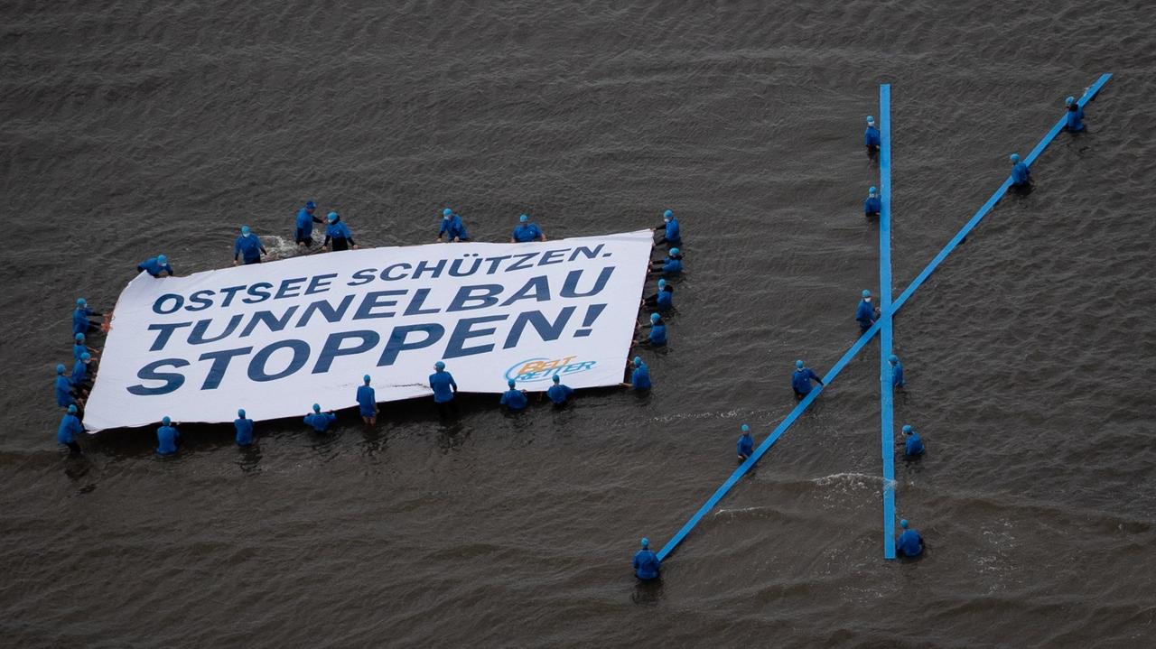 Aktivisten der Protestaktion "Beltretter" halten ein Banner mit dem Text "Ostsee schützen. Tunnelbau stoppen!", während sie im Ostseewasser stehen. Aktivisten der Protestaktion "Beltretter" halten ein Banner mit dem Text "Ostsee schützen. Tunnelbau stoppen!", während sie im Ostseewasser stehen.