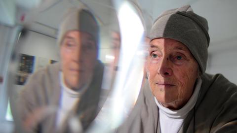 Die Künstlerin Mary Bauermeister trägt ein Tuch um den Kopf, sie betrachtet eines ihrer Kristall-Objekte. Die Ausstellung, in dem ihr Gesicht auch zu sehen ist.