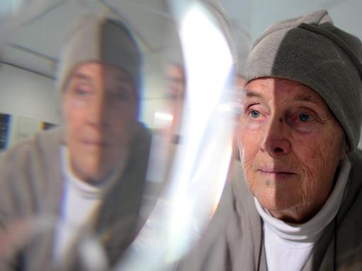 Die Künstlerin Mary Bauermeister trägt ein Tuch um den Kopf, sie betrachtet eines ihrer Kristall-Objekte. Die Ausstellung, in dem ihr Gesicht auch zu sehen ist.