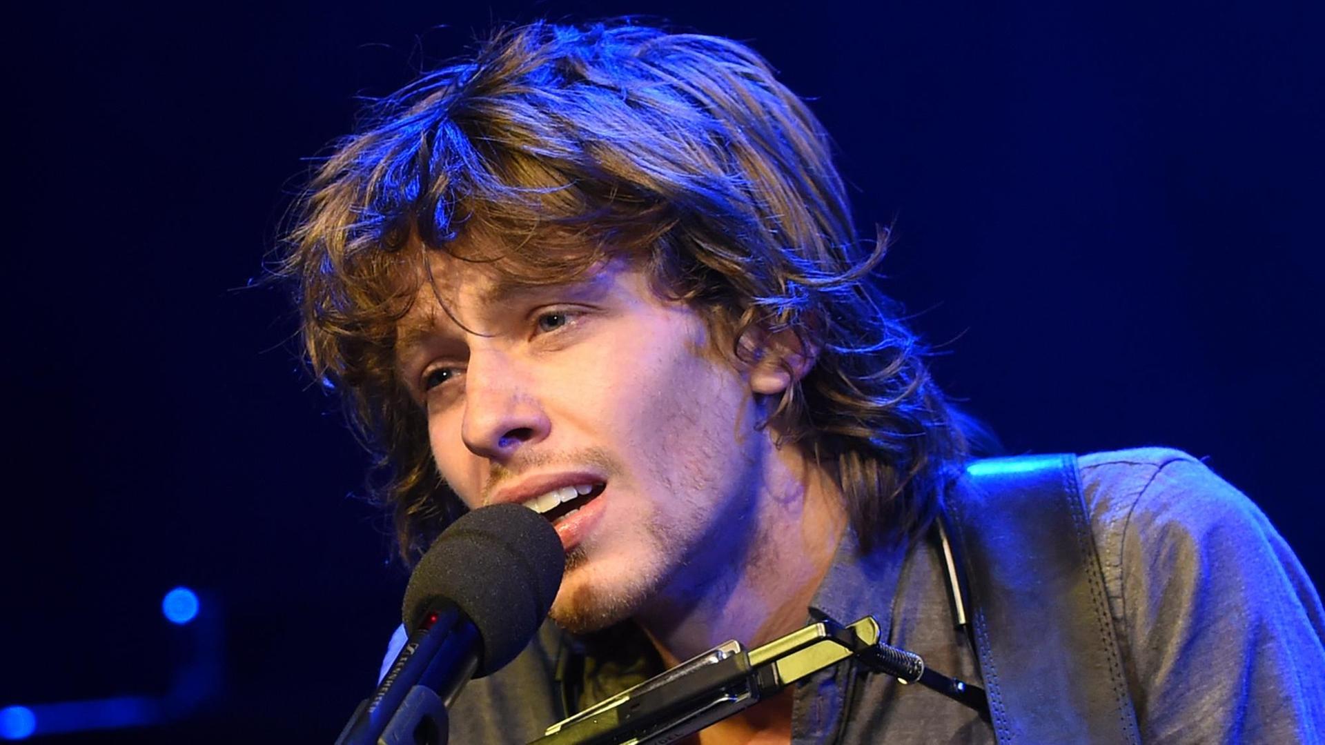 Ein junger Mann mit buschigem Haar singt mit leidenschaftlichem Ausdruck in ein Mikrofon. Ein junger Mann mit buschigem Haar singt mit leidenschaftlichem Ausdruck in ein Mikrofon.