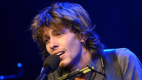 Ein junger Mann mit buschigem Haar singt mit leidenschaftlichem Ausdruck in ein Mikrofon.