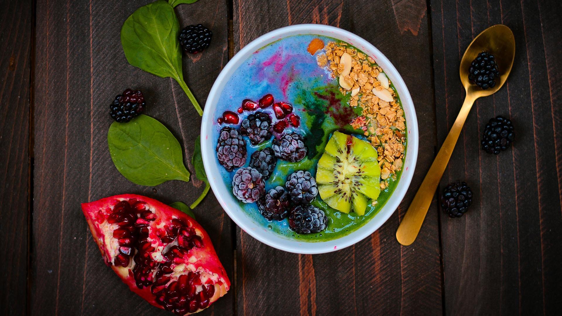 Eine Müslischale mit einer aufgeschnittenen Kiwi, Brombeeren, Granatapfelkernen und Müsli. Die Farben laufen ineinander. Daneben liegen ein goldener Löffel, ein Stück Granatapfel, zwei Spinatblätter und einige Brombeeren auf einem dunklen Tisch. Eine Müslischale mit einer aufgeschnittenen Kiwi, Brombeeren, Granatapfelkernen und Müsli. Die Farben laufen ineinander. Daneben liegen ein goldener Löffel, ein Stück Granatapfel, zwei Spinatblätter und einige Brombeeren auf einem dunklen Tisch.