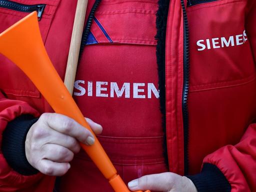 Nahaufnahme des Oberkörpers eines protestierenden Siemens-Mitarbeiters in roter Jacke mit dem Konzernlogo und Tröte in der Hand