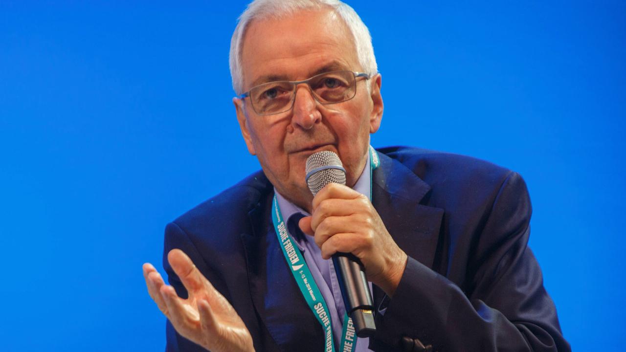 Klaus Töpfer sitzt vor einer blauen Wand und spricht in ein Mikrophon, dabei hebt er leicht eine Hand Klaus Töpfer sitzt vor einer blauen Wand und spricht in ein Mikrophon, dabei hebt er leicht eine Hand