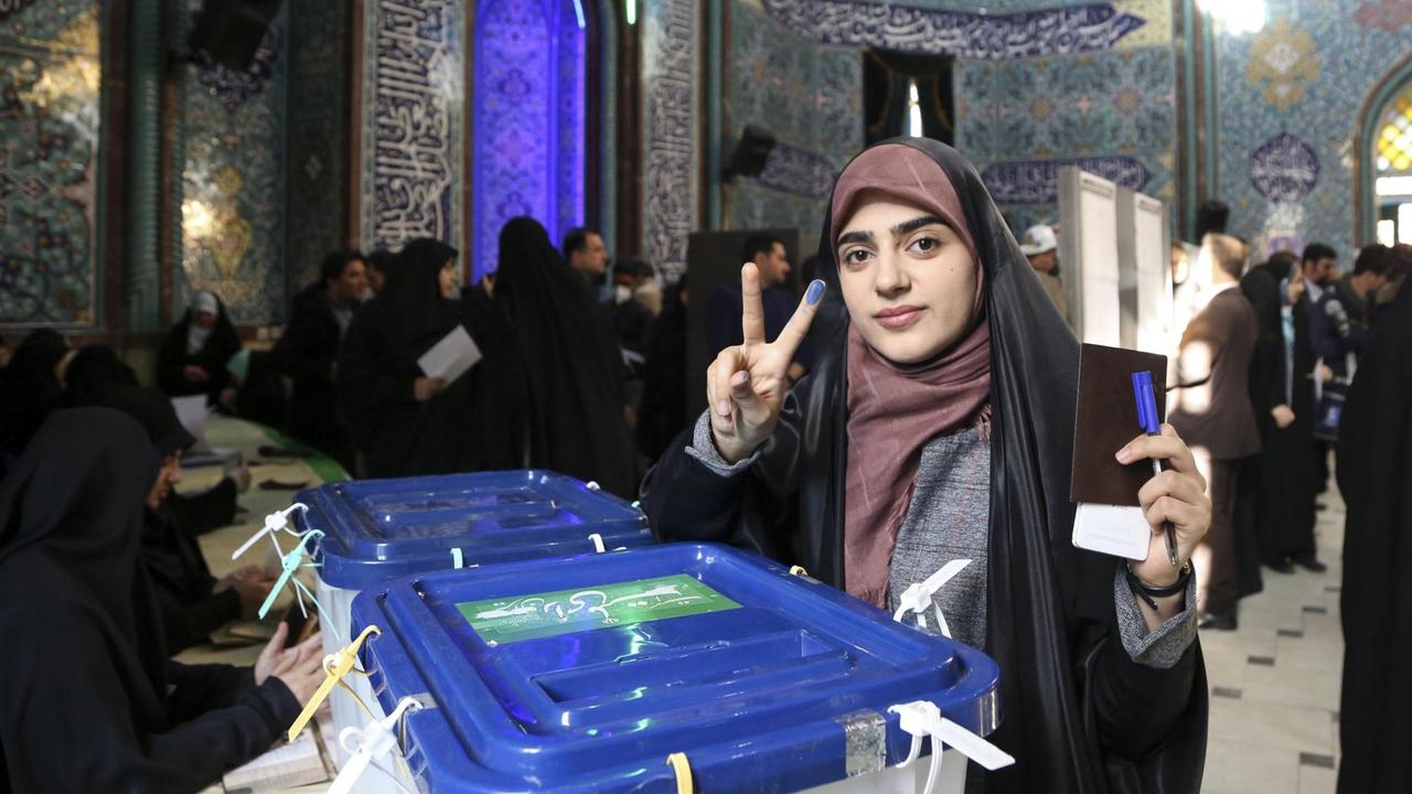 Eine Frau an den Wahlurnen macht das Victory-Zeichen. In Zeigefinger ist mit blauer Tinte eingefärbt.