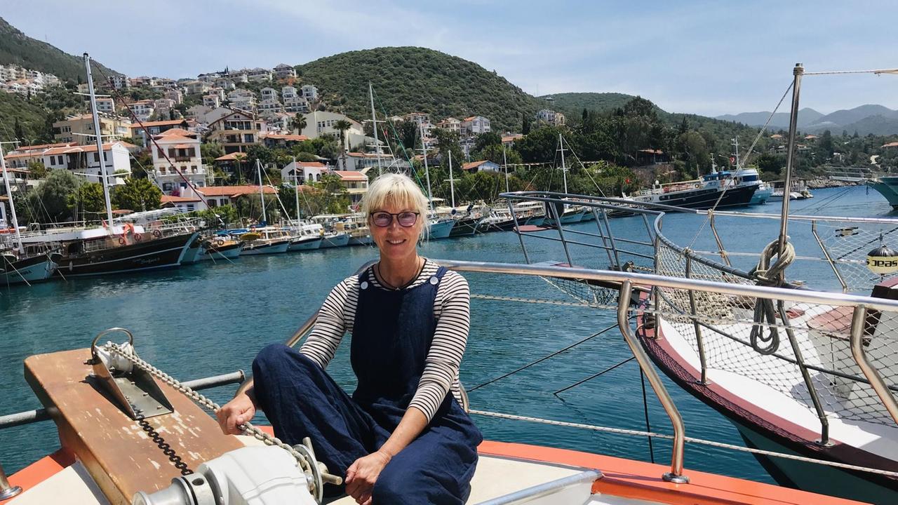 Eine blonde Frau in dunkelblauer Latzhose und mit Brille sitzt auf einem Boot. Im Hintergrund das Meer und die andere Seite des Hafens mit vielen Schiffchen. Eine blonde Frau in dunkelblauer Latzhose und mit Brille sitzt auf einem Boot. Im Hintergrund das Meer und die andere Seite des Hafens mit vielen Schiffchen.