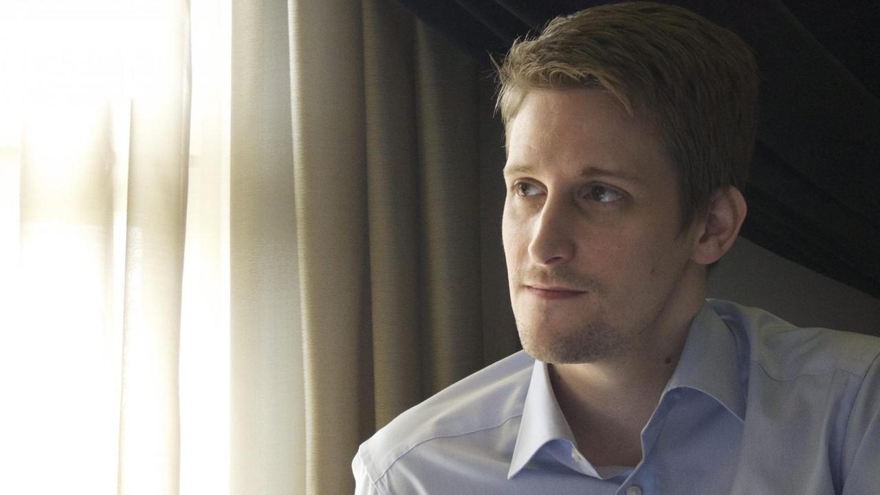 Snowden sitzt mit melancholischem Blick vor einem Fenster mit zugezogenen Gardinen.