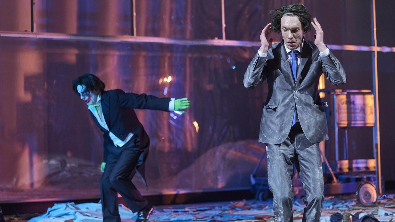 Szenenfoto: Ein Mann im Anzug und mit zerzausten Haaren hebt die Hände Richtung Kopf. Neben ihm steht ein Mann, der aussieht wie die Comic-Figur "Joker". Verwüstung auf der Bühne. Szenenfoto: Ein Mann im Anzug und mit zerzausten Haaren hebt die Hände Richtung Kopf. Neben ihm steht ein Mann, der aussieht wie die Comic-Figur "Joker". Verwüstung auf der Bühne.