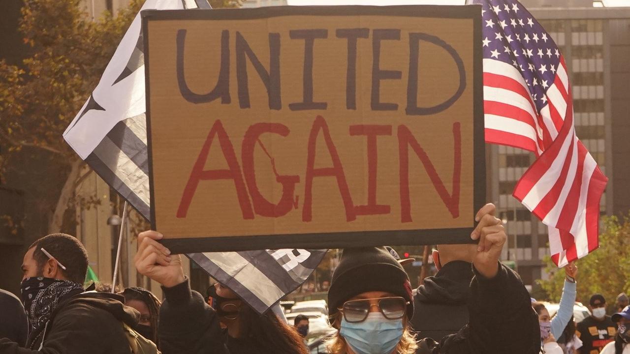 Eine Frau mit Mund-Nasen-Schutz hält ein Schild hoch, worauf steht: "United again", im Hintergrund eine amerikanische Fahne
