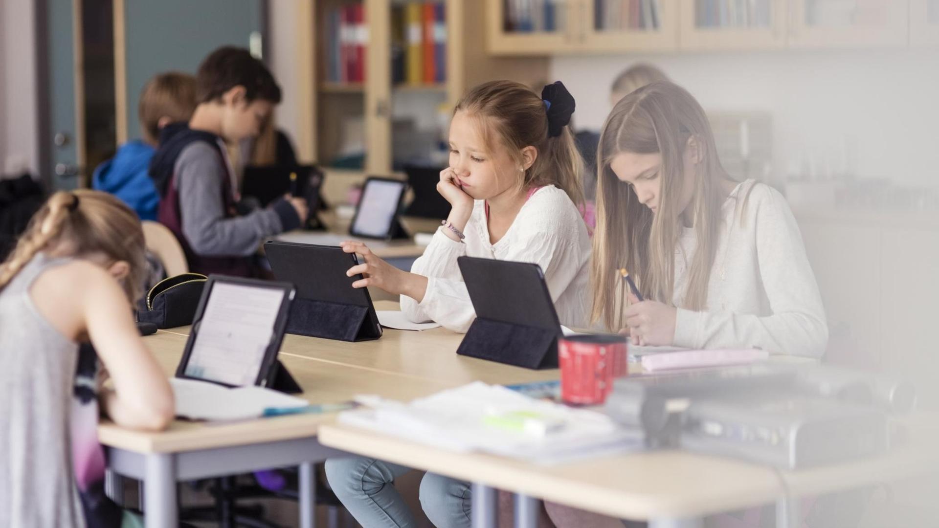 In einem Klassenzimmer sitzen Kinder die an Tablets arbeiten. In einem Klassenzimmer sitzen Kinder die an Tablets arbeiten.