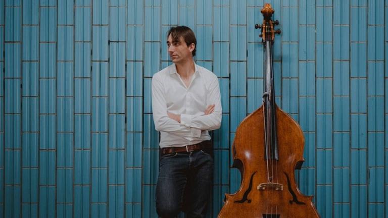 In Jeans und mit weißem Hemd sowie seinem Kontrabass steht der Musiker vor einer blauen Kachelwand. In Jeans und mit weißem Hemd sowie seinem Kontrabass steht der Musiker vor einer blauen Kachelwand.