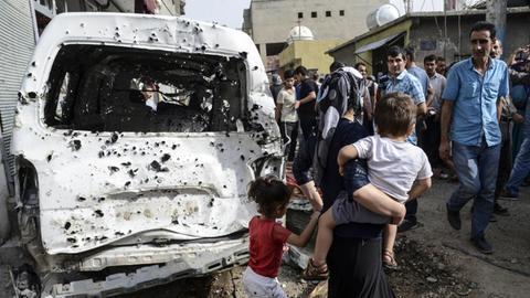 Zu sehen ist ein Bild aus der Stadt Cizre im Südosten der Türkei, im Vordergrund ein zerstörtes Auto, rechterhand eine Mutter mit einem Kind.