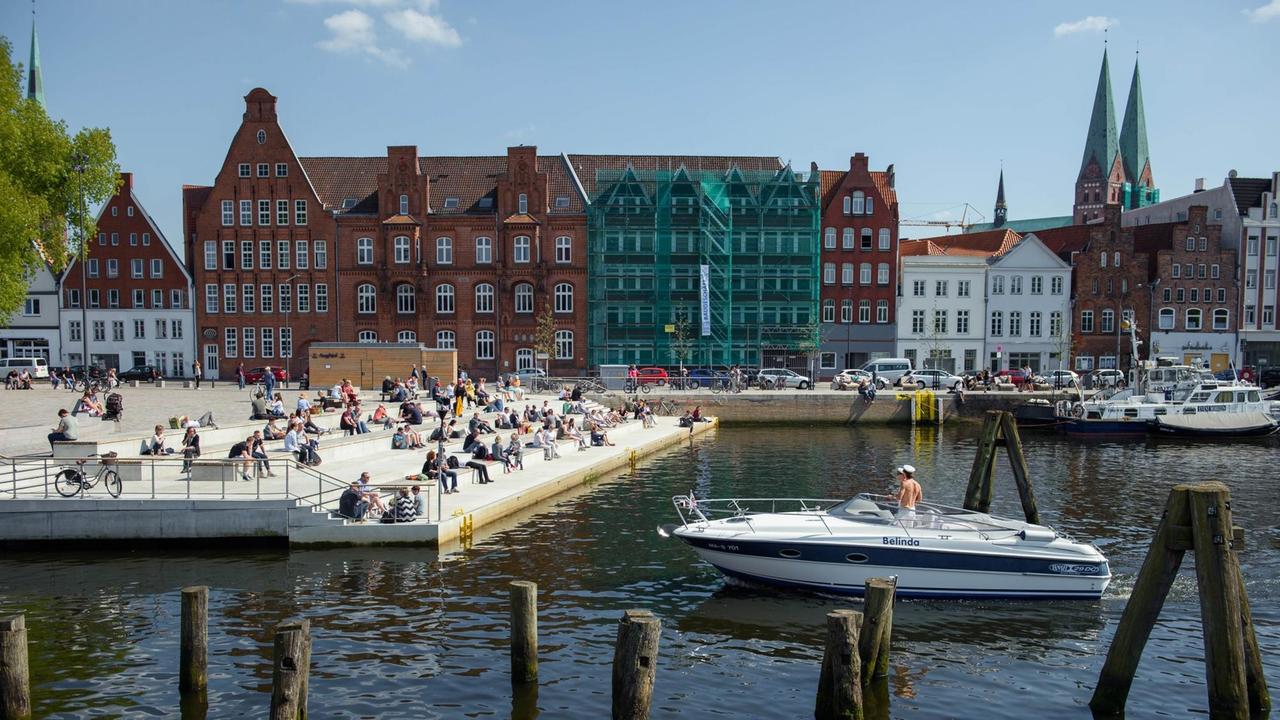 In Lübeck sitzen bei Sonnenschein viele Menschen auf den Stufen des Drehbrückplatz am Ufer der Trave, während zwei Männer in einem Boot vorbeifahren. IM Hintergrund ist die Lübecker Altstadt zu sehen.