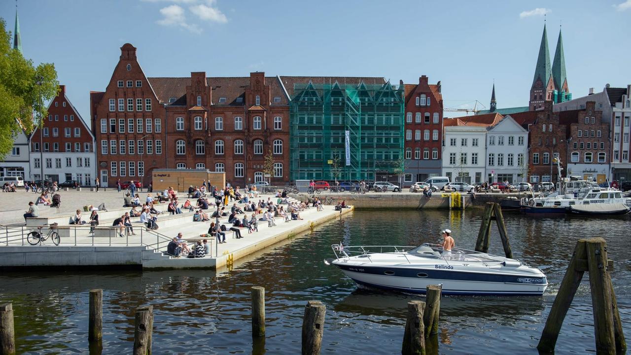 In Lübeck sitzen bei Sonnenschein viele Menschen auf den Stufen des Drehbrückplatz am Ufer der Trave, während zwei Männer in einem Boot vorbeifahren. IM Hintergrund ist die Lübecker Altstadt zu sehen. In Lübeck sitzen bei Sonnenschein viele Menschen auf den Stufen des Drehbrückplatz am Ufer der Trave, während zwei Männer in einem Boot vorbeifahren. IM Hintergrund ist die Lübecker Altstadt zu sehen.