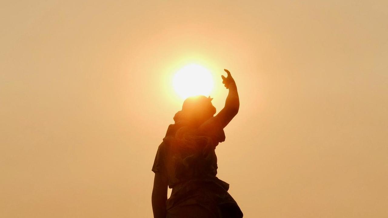 Die untergehende Sonne berüht die Silhouette einer Statue, mit erhobener rechter Hand.