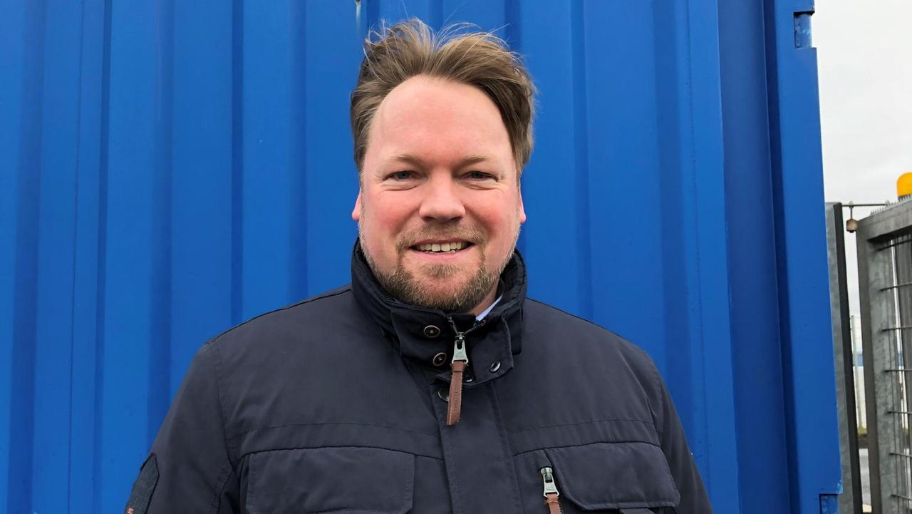 Der Wind weht Oliver Kubartzky durch die Haare. Er steht vor der Wand eines blauen Containers. Der Wind weht Oliver Kubartzky durch die Haare. Er steht vor der Wand eines blauen Containers.