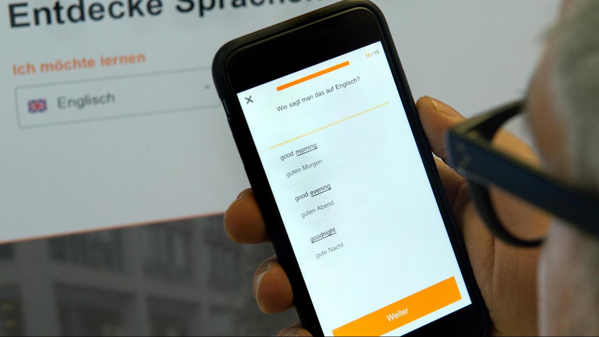 Ein Mann hält ein Smartphone mit der Applikation (App) «Babbel» zum Erlernen einer Fremdsprache vor einen Monitor. Ein Mann hält ein Smartphone mit der Applikation (App) «Babbel» zum Erlernen einer Fremdsprache vor einen Monitor.