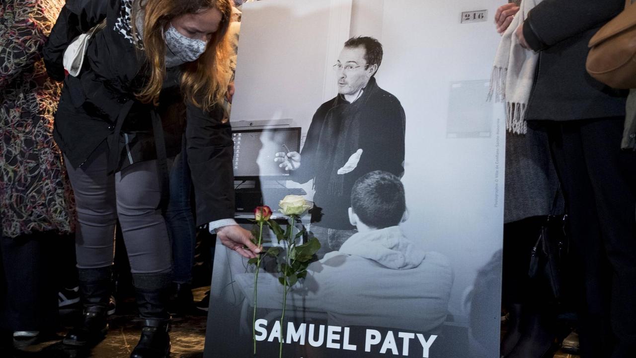 Das Foto zeigt Menschen auf einer Demonstration für die Meinungsfreiheit und gegen islamistischen Terror, die Blumen vor einem Bild des ermordeten Lehrers Samuel Paty ablegen.  