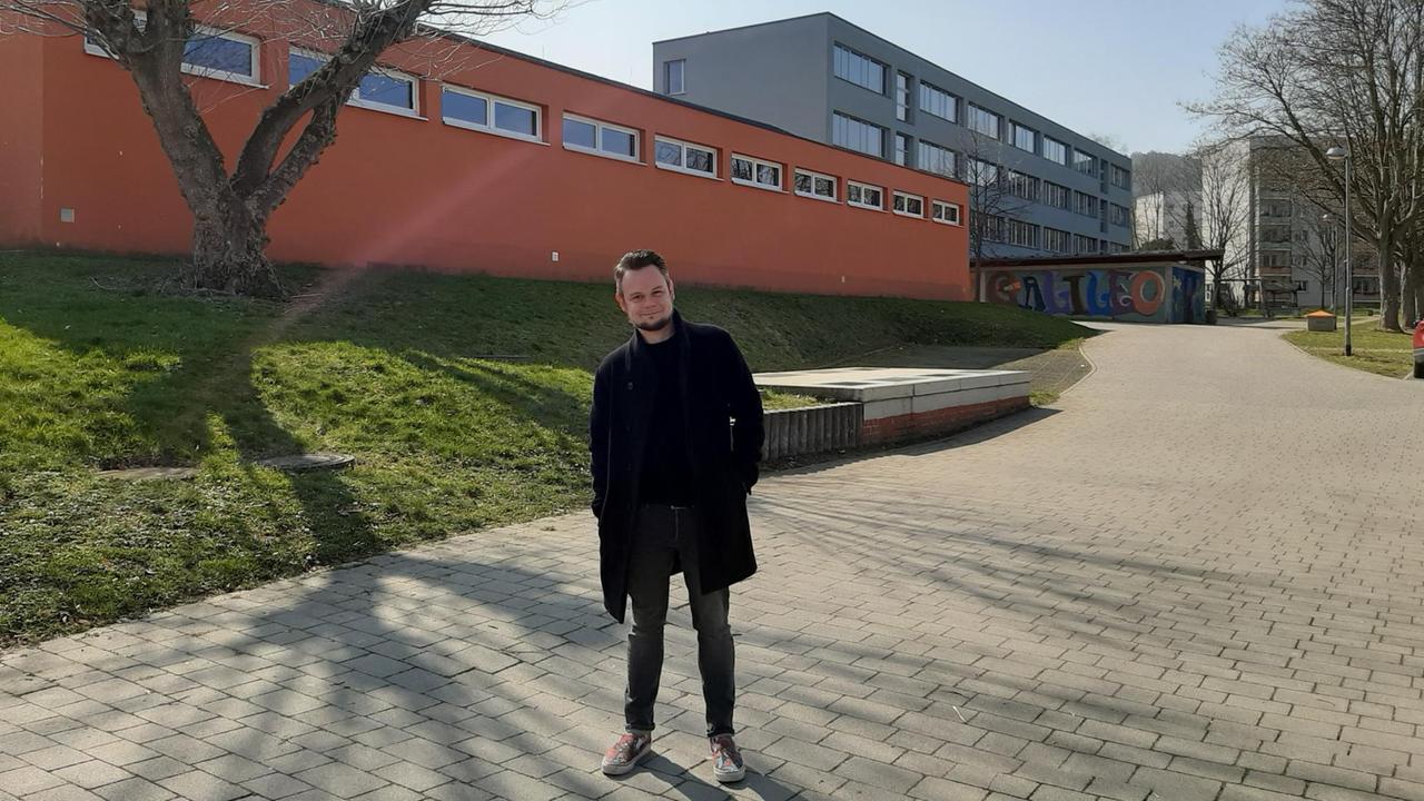 Ein junger Mann steht vor einem Schulgebäude.
